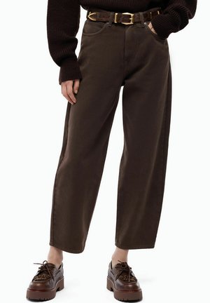 Donna che indossa pantaloni marrone scuro a gamba larga, una cintura marrone con fibbia dorata, un maglione marrone scuro lavorato a maglia e scarpe marroni chunky con lacci.