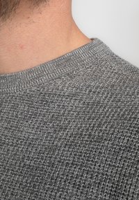 Grauer, gestrickter Pullover aus einem texturierten, atmungsaktiven Stoff. Eine Nahaufnahme zeigt einen gerippten Kragen und ein weiches, mittelschweres Gewebe.