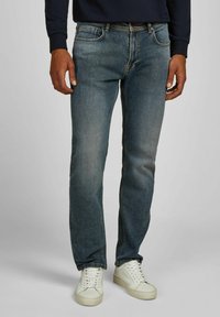 Homme portant un jean bleu délavé, des baskets blanches et une chemise à manches longues noire, se tenant devant un fond gris clair uni.