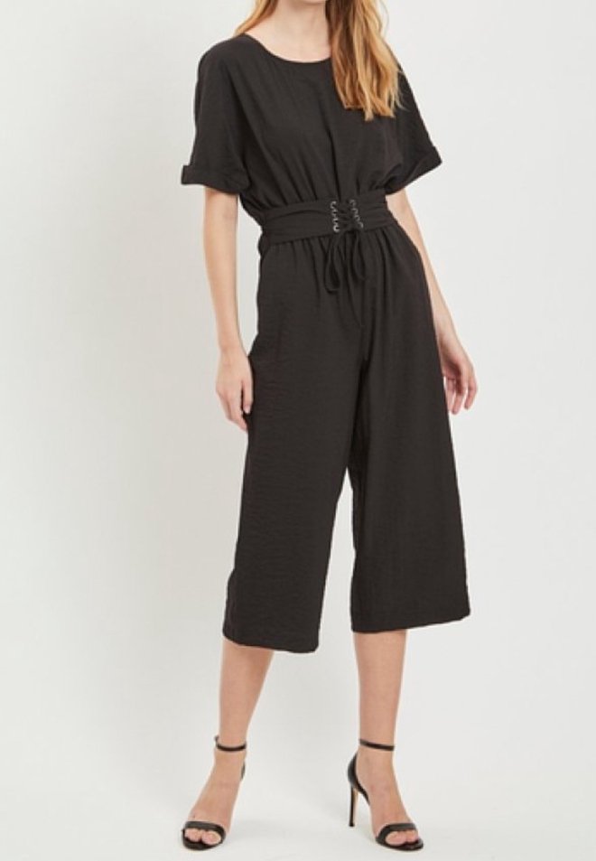 Femme portant une combinaison ample noire à manches courtes, taille cintrée et jambes larges raccourcies, associée à des sandales noires à talons hauts.