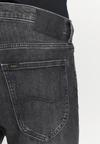 Pantalones vaqueros de mezclilla negra con un bolsillo trasero, con la etiqueta de la marca Lee cosida encima del bolsillo y un parche en la cinturilla sobre un fondo blanco.