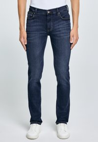 Mężczyzna ubrany w ciemnoniebieskie, dopasowane jeansy z pięcioma kieszeniami, białe trampki i biały top, stojący na tle jednolitego jasnego tła.