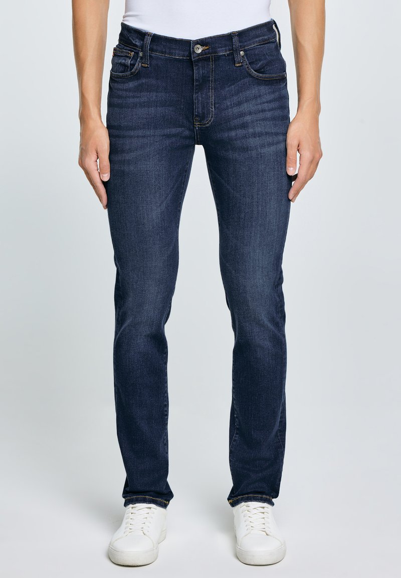 Mężczyzna ubrany w ciemnoniebieskie, dopasowane jeansy z pięcioma kieszeniami, białe trampki i biały top, stojący na tle jednolitego jasnego tła.