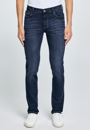 Muž nosící tmavě modré slim fit džíny s pěti kapsami, bílé tenisky a bílý top, stojící před jednobarevným světlým pozadím.