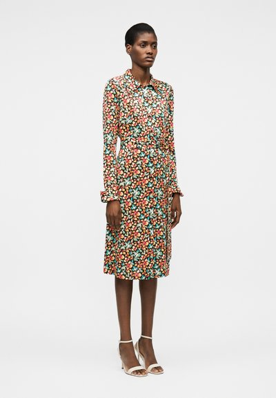 Γυναίκα που στέκεται, φορώντας μακρυμάνικο, με γιακά, φόρεμα μήκους μέχρι το γόνατο με πολύχρωμο ανθηρό μοτίβο και λευκές ψηλοτάκουνες σανδάλες σε απλό φόντο.