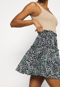 Marineblauwe bloemenrok met groene en roze patronen, voorzien van een rimpelrand en een elastische tailleband, gecombineerd met een beige geribd bodysuit.