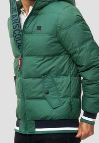 Giacca a vento verde con cappuccio, caratterizzata da un design trapuntato, fodera interna, zip frontale e due tasche laterali con bottoni. Orlo a coste con righe.