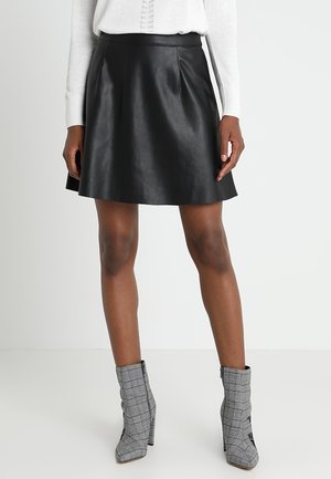 Femme portant une jupe en cuir noir trapèze, un pull blanc, et des bottines grises à carreaux avec talons carrés, debout devant un fond uni.