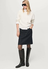 Maglione bianco a trecce, gonna di jeans blu e stivali neri fino al ginocchio. La modella indossa occhiali da sole neri oversize, con un'acconciatura elegante.