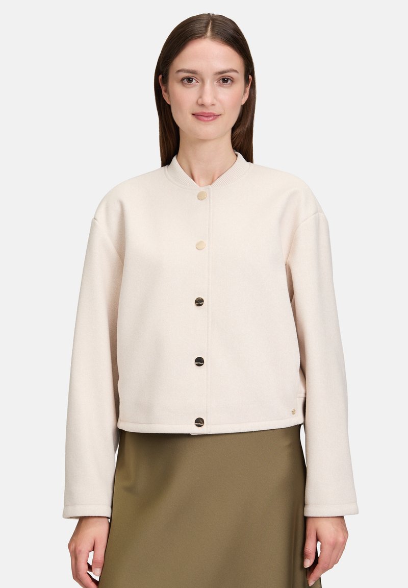 Krótka, przycięta kremowa kurtka wykonana z miękkiego materiału, z okrągłym dekoltem i pięcioma metalowymi guzikami z przodu. Długie rękawy i minimalistyczny design.