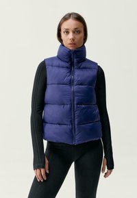 Temno modra napihnjena brezrokavnik z visokim ovratnikom in sprednjim zadrgo. Ponuja horizontalno prešito teksturo in gladko, sijočo površino.