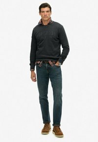 Hombre de pie con sudadera negra sobre una camisa de cuadros rojos, jeans azules, botas de cuero marrón y reloj, posando frente a un fondo blanco.