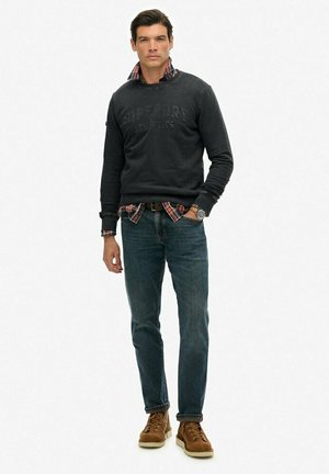 Man die staat met een zwart sweatshirt over een rood geruit overhemd, blauwe jeans, bruin leren laarzen en een horloge, poserend tegen een witte achtergrond.