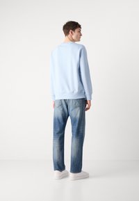 Light blue sweatshirt with ribbed cuffs and hem, paired with light blue, faded jeans. Model wears white sneakers and stands against a neutral backdrop.