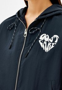 Osoba w granatowej bluzie z zamkiem błyskawicznym, z białymi sznurkami i sercowym napisem "Nie Obchodzi Mnie" na piersi.