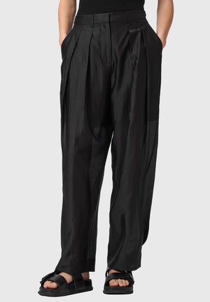 Luźne czarne plisowane spodnie noszone z czarnymi sandałami z odkrytymi palcami i rękami w kieszeniach, widoczne od pasa do stóp.