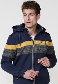 Sudadera con cremallera en tonos marino y amarillos, con una franja gris texturizada, bolsillos delanteros y capucha con cordones. Logo de acento en la manga.