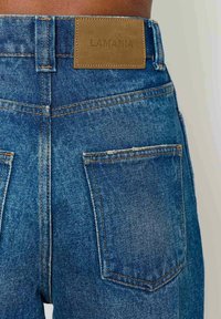 Džinsai su mėlyna spalva, turintys odinę etiketę su užrašu "LAMANIA" ir kontrastinėmis geltonomis siūlėmis. Matomos standartinės kišenės ir siuvimo detalės.