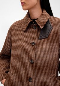 Casaco castanho aos quadrados com gola estruturada, detalhe em couro na gola, cinco botões pretos e dois bolsos laterais. Tecido texturizado.