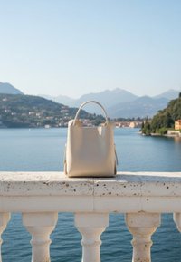 Κρεμ τσάντα tote ξαπλωμένη σε πέτρινο μπαλκόνι με θέα σε μια ήρεμη λίμνη με μακρινούς βουνά και σπίτια κάτω από καθαρό ουρανό.