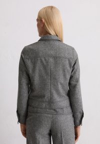 Giacca grigia in misto lana con un colletto strutturato, vestibilità sartoriale e dettagli di cucitura sottili, vista da dietro, abbinata a pantaloni su misura.