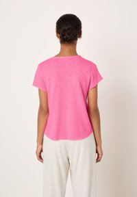 Ženska s temnimi lasmi povezanimi v čop, oblečena v kratkoročno svetlo roza majico in svetlo sive hlače, stoji obrnjena proti enobarvnemu belemu ozadju.