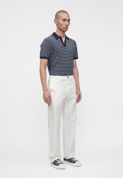 Homme debout, portant un polo à rayures bleu marine et blanc, un pantalon blanc et des baskets bleu marine avec des semelles et des lacets blancs, sur un fond uni.