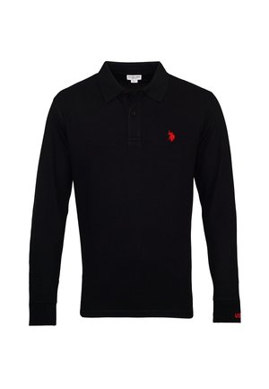 Homme portant un polo noir à manches longues avec un logo rouge, un jean bleu et des baskets bleu marine, debout devant un fond uni.