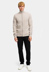 Svijetlo bež rebrasti džemper na patentni zatvarač s visokim ovratnikom, uparen s crnim hlačama i bijelim tenisicama, stojeći ispred neutralne pozadine.