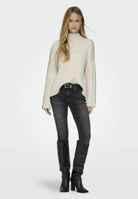 Maglione lavorato a maglia di colore crema con texture a coste e dettagli neri sui lati, abbinato a jeans skinny neri e stivali neri con tacco al ginocchio.