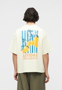 Άτομο με κοντά σκούρα μαλλιά που φορά ένα κρεμ t-shirt Adidas Originals με μπλε ρίγες, κίτρινα φρούτα και πορτοκαλί κείμενο στην πλάτη.