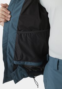 Veste imperméable en teal avec une poche en filet noire à l'intérieur. Elle présente une texture lisse, un ourlet ajustable avec cordon de serrage et une fermeture à bouton-pression.
