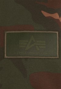 Zöld terepmintás anyag, egy téglalap alakú zöld folttal, amelyen az "ALPHA INDUSTRIES" felirat és egy stilizált logó található a közepén.