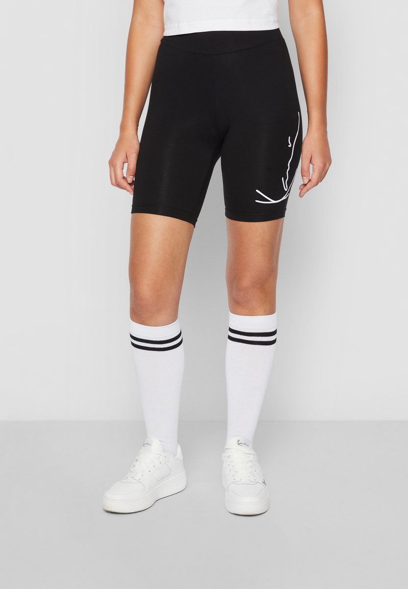 Person som bär svarta cykelshorts med vit text på sidan, vita knästrumpor med svarta ränder och vita sneakers.