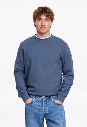 Jonge man met krullend haar, die een blauwe trui en lichtblauwe spijkerbroek draagt, staat met handen in de zakken tegen een witte achtergrond.