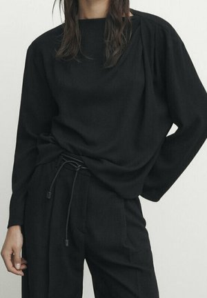 Person wearing loose black long-sleeve top and matching black pants with drawstring waist, standing against a plain light background.