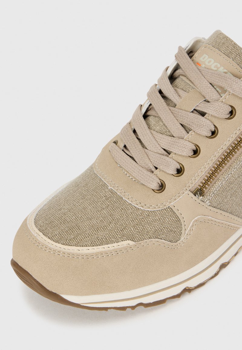 Zapatilla beige con parte superior de lona y ante, puntera redonda, cordones planos, cremallera lateral y suela de goma con acentos en blanco y marrón.