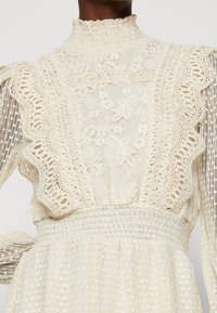 En spetsklänning i krämfärg med hög hals, intrikata blommönster, volangdetaljer och textil med vertikala ränder och prickar.