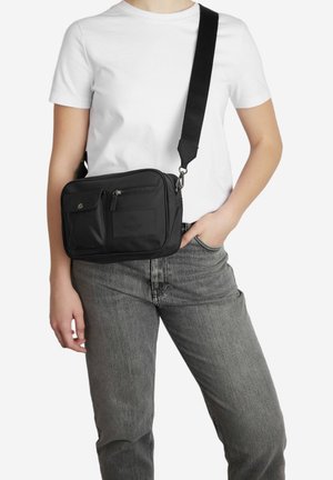 Czarna nylonowa torba na ramię z zamykaną główną przegrodą, przednią kieszenią i regulowanym paskiem, prezentowana na modelu w białej koszulce.