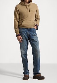 Felpa marrone con tasca frontale, jeans blu e scarpe marroni. La felpa ha un cordino e un logo sul petto; i jeans sono sbiaditi.