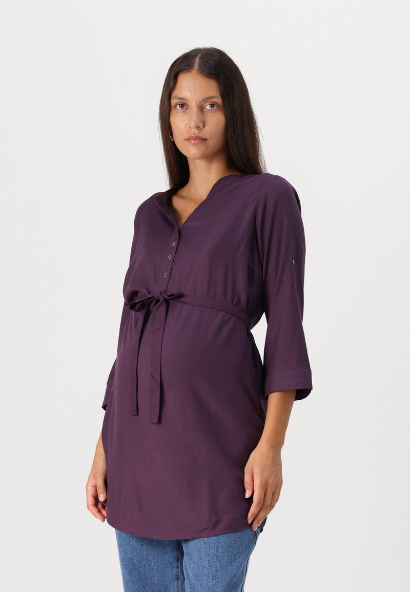 Tunic viola per maternità con scollo a V, maniche a tre quarti e una cintura frontale. Realizzata in tessuto morbido, offre una vestibilità comoda.