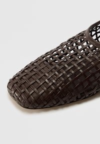 Zapato marrón tejido tipo slip-on con un diseño abierto, que presenta una puntera cuadrada y una textura suave. Fabricado con material flexible y un patrón detallado.