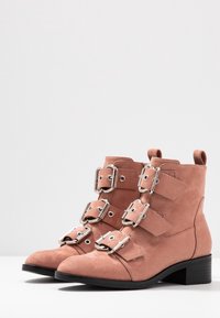 Růžové semišové kotníkové boty se třemi přezkami, stříbrnými detaily a nízkým blokovým podpatkem. Strukturovaný povrch a zaoblený tvar špičky.