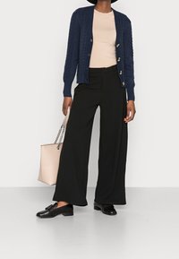 Marinblå stickad cardigan, ljusbeige topp, svarta vida byxor, svarta loafers, håller en rosa tygväska med kedjehandtag. Enkel och funktionell design.