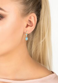 Pendientes de oro con forma de gota que presentan una piedra preciosa en tono azul claro en forma de lágrima, acentuada por una pequeña piedra redonda justo encima. Diseño detallado y delicado.