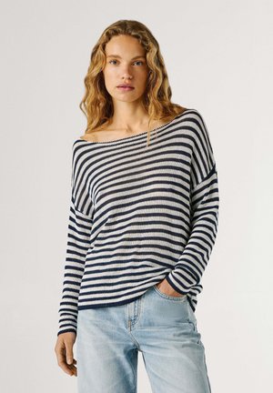 Jeune femme aux cheveux blonds ondulés portant un pull à rayures bleu marine et blanc, épaules découvertes, et un jean bleu clair, main dans la poche, fond neutre.