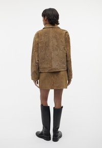 Giacca in suede marrone con decorazioni metalliche, abbinata a una gonna coordinata e stivali neri fino al ginocchio. Texture liscia e design sartoriale.