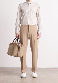 Väriltään vaaleanruskeat housut ja vaalea raidallinen nappipaita, kädessään beigeä ruskein yksityiskohdin varustettua laukkua. Jalassa yksinkertaiset valkoiset lenkkarit.