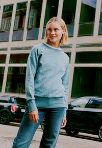 Felpa azzurra con texture a macchie, polsini e orlo a costine. Presenta tre bottoni decorativi sulla spalla sinistra.