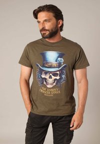 T-shirt en coton vert olive présentant un grand motif de crâne avec un haut-de-forme bleu et une rose, texte imprimé : "LE STYLE INFINI DU DANDY."